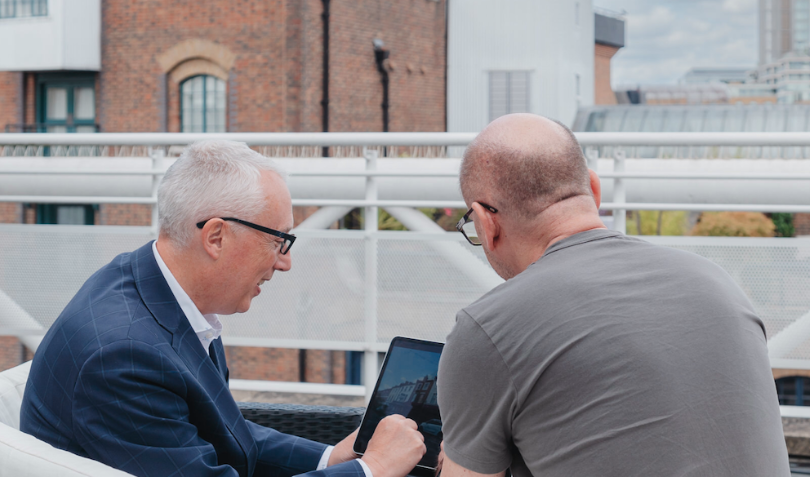 Two men are sitting on a sofa on a rooftop overlooking London. They are both looking at a screen.