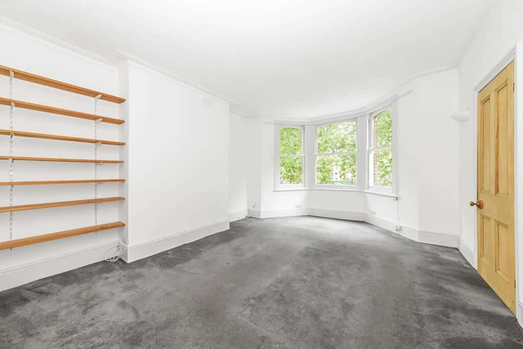 An empty room with a large bay window and empty wooden shelves. The walls are white with grey carpet.