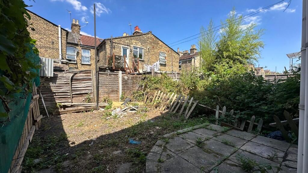 A small garden with a patio which has lots of weeds growing through the cracks. Theres a damaged fence and unmaintained greenery. There also seems to be rubbish left on the grass.