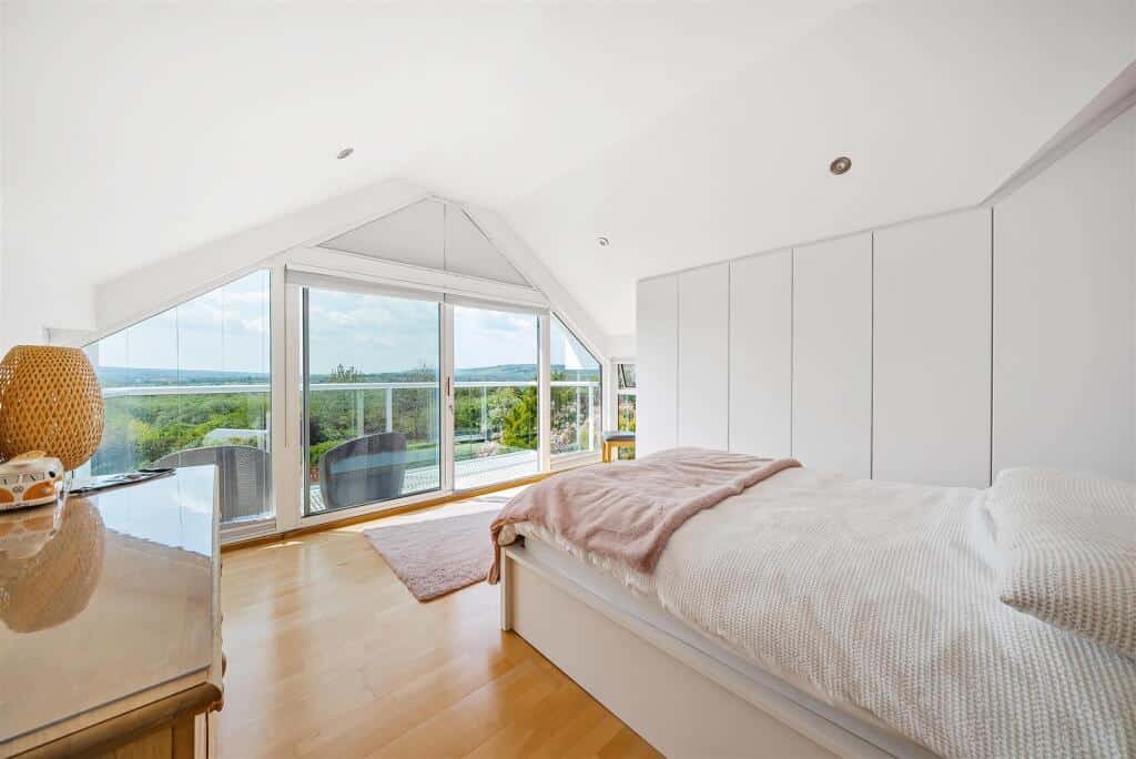 A larger room with white walls, wooden floors and a sloped ceiling. One wall has floor to ceiling windows with a glass door that leads out onto a balcony and another has a inbuilt wardrobe.