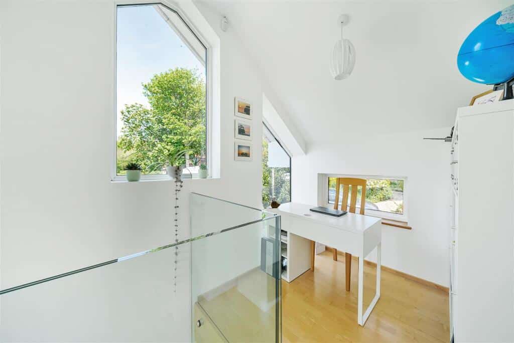 A small alcove office near the stairs with large windows and glass railings. Theres a white desk with a laptop.