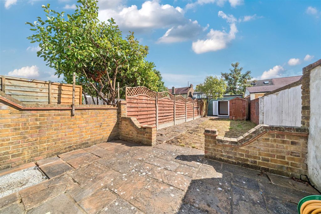 A long narrow back garden with a clean patio and patio wall. At the end of the garden theres a tree and a shed.
