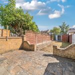 A long narrow back garden with a clean patio and patio wall. At the end of the garden theres a tree and a shed.