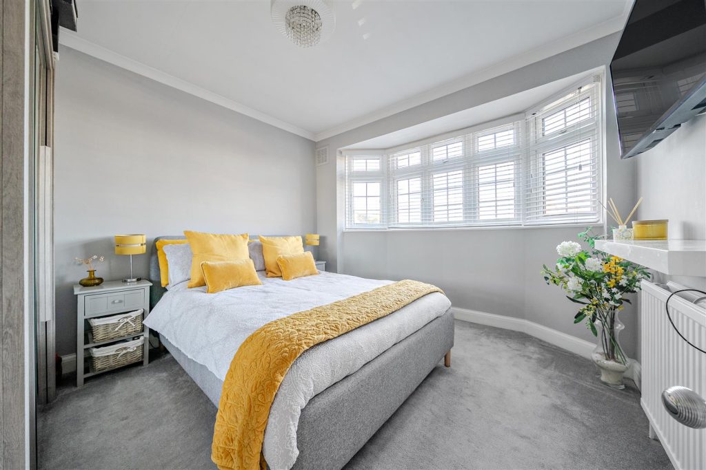 A double-size master bedroom shows a bay window. Sunlight brightens the space. A well-made bed with yellow accent cushions and blankets add a pop of colour to the smart space.
