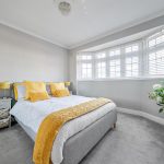 A double-size master bedroom shows a bay window. Sunlight brightens the space. A well-made bed with yellow accent cushions and blankets add a pop of colour to the smart space.