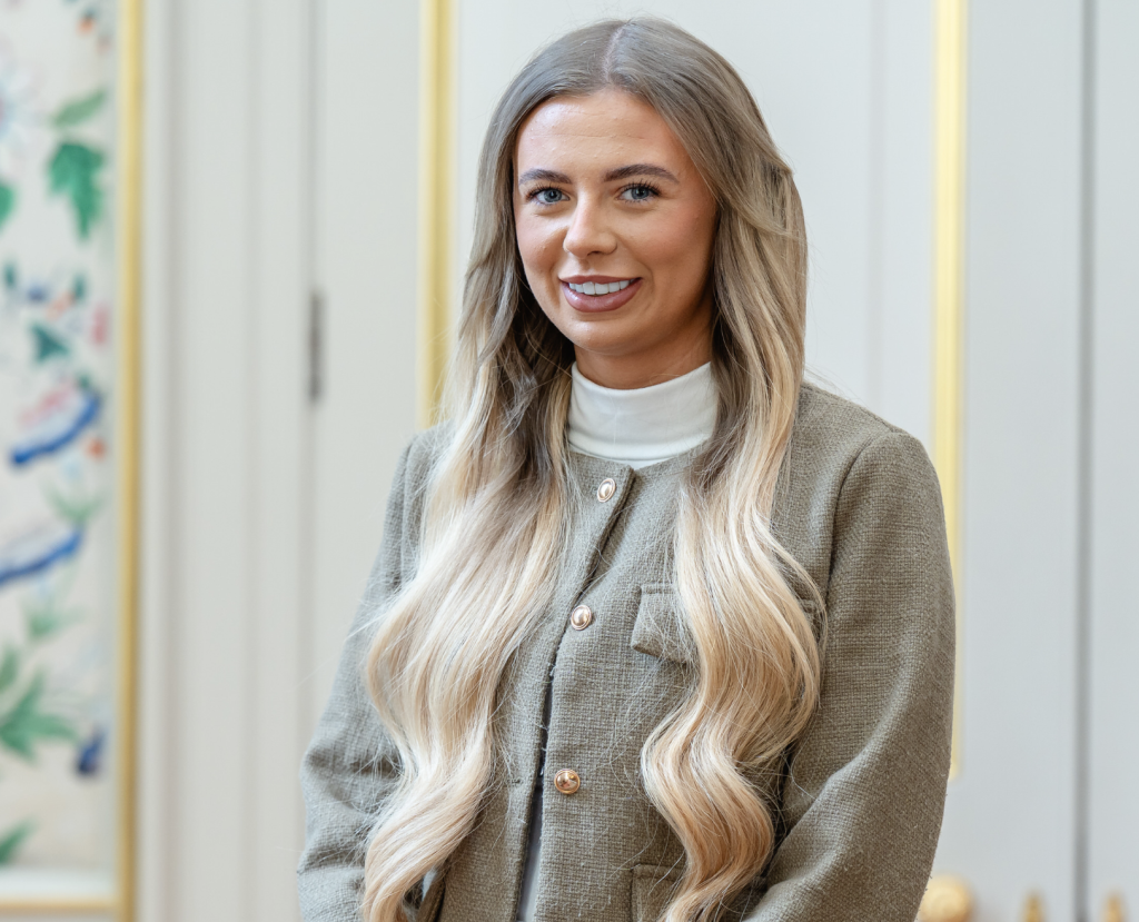 Poppy smiles into the camera. She is in front of a decadent background and wearing a white high neck jumper and a beige blazer.