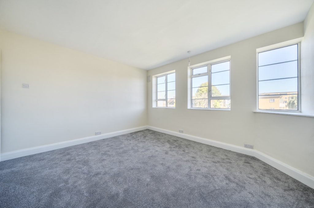 A large room with clean white walls and a new dark grey carpet. The room is well lit thanks to the three large windows.