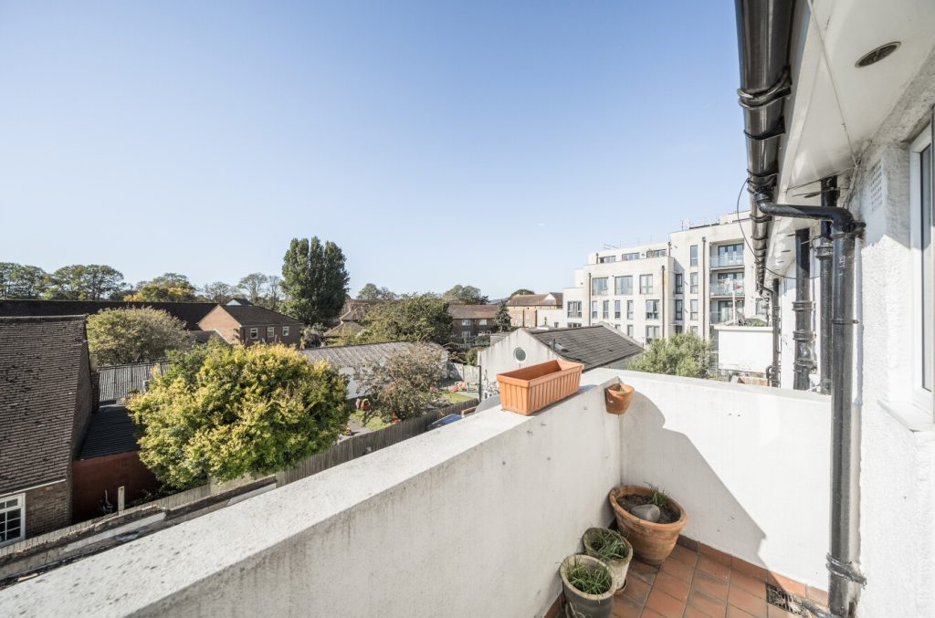 The balcony is a cosy, brightly-lit area with a few plant pots. The walls are white concrete and the floors are terracotta coloured tiles.
