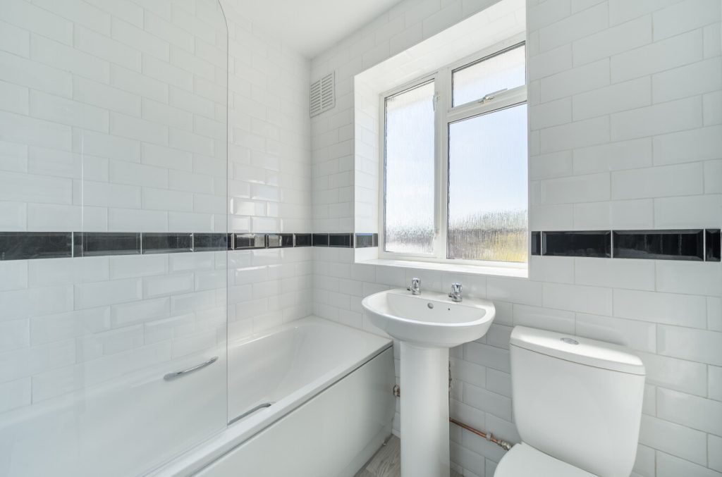 Bathroom has a shower over the bath with a clean glass screen. There's a white sink next to the bath and a white toilet next to that. Walls are white tiled with a black border in the middle.