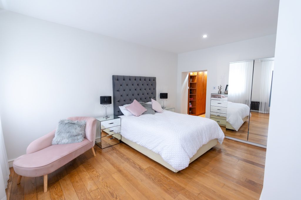 The master bedroom currently holds a large king-size bed and additional chaise lounge, a free standing mirror, and chest of drawers. At the far side of the room, an oak door is open to expose the dressing room.