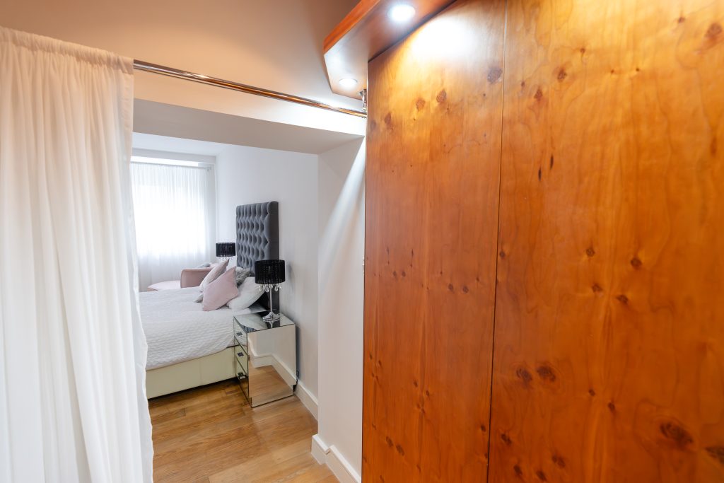 Peeking into the Master Bedroom from the Dressing Room, you can see the head of the large bed – bordered each side by mirrored nightstands. Behind the bed, a large sash window is covered by a delicate, sheer curtain, letting in lots of light.