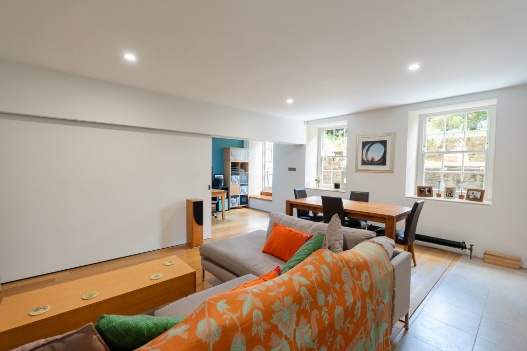 A large living room features a large, "L-shape" sofa in front of a six-seater dining table. Sash windows at the end of the room give lots of light. On the far side of the room, the media wall is opened slightly to expose the second bedroom – currently used as a home office.