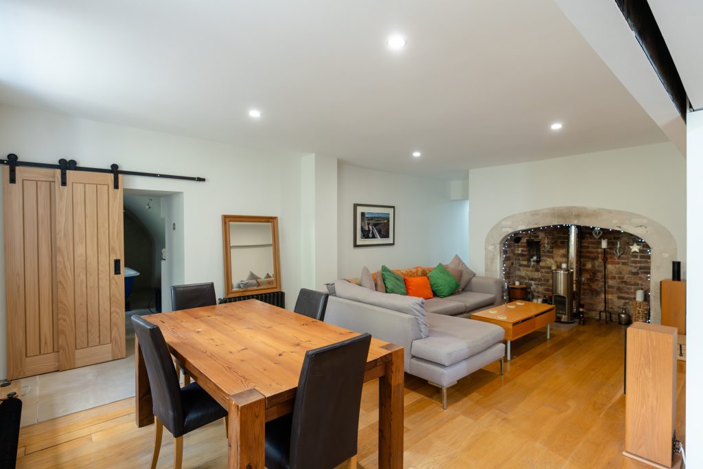 Taken from the room's windows, you can see the large dining table and L-shaped sofa stretching out into the expanse of the living area. On the left side of the room, a barn door hovers, half-slid open. On the other end of the room, you can glimpse the connection to the kitchen. In the farest corner sits the large, original fireplace.
