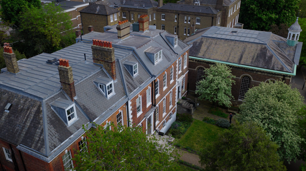 Spencer House is a classical era listed building. It is pictured from the air, showing a pitched, grey slate-covered roof lined with five dormer windows. Its front is split into five bays, each are red brick with stone dressings. Image shows five storeys to the building.