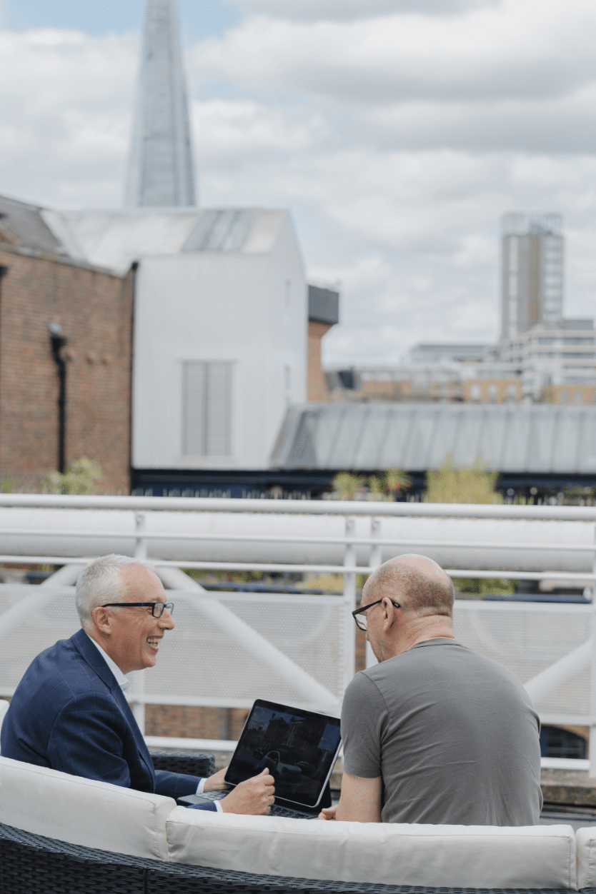 Two men are discussing the contents of a laptop screen whilst overlooking London milestones